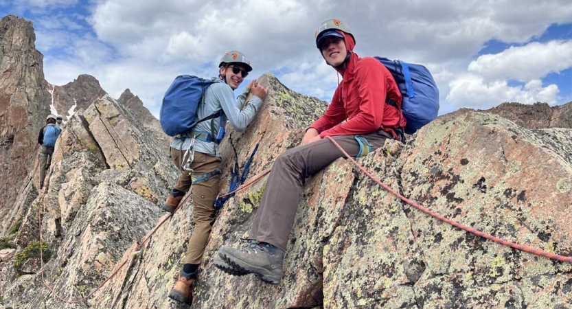 a person wearing safety gear sits on a rock, while another wearing safety gear climbs up to be beside them